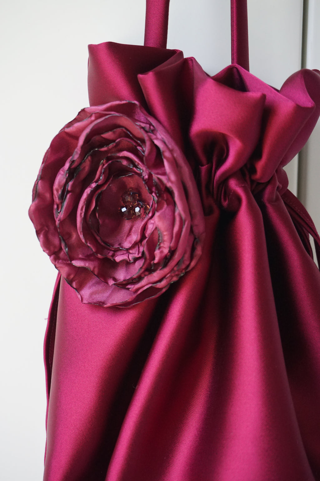 A close-up of a deep magenta satin drawstring bag featuring a large matching fabric rose embellishment on the front.