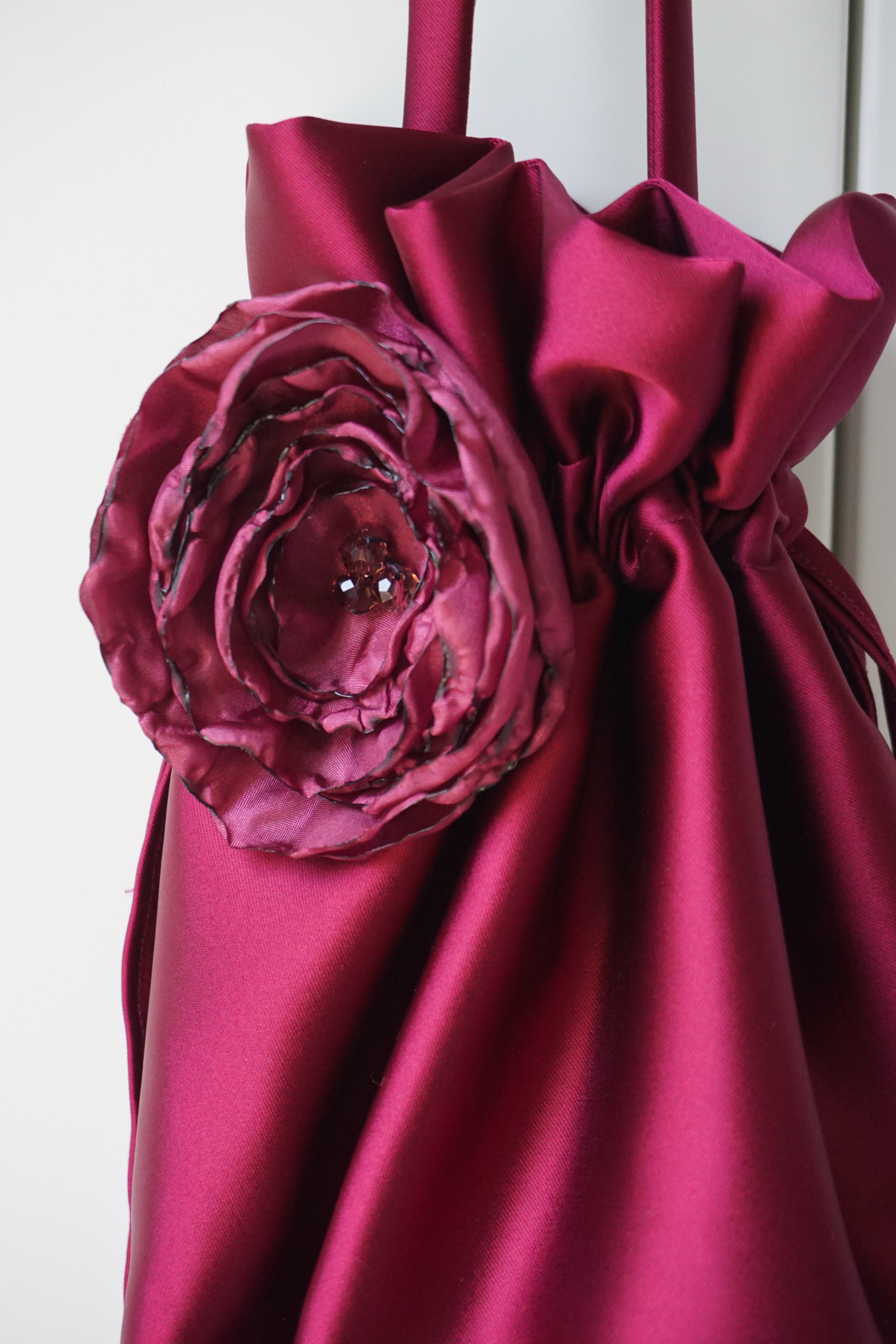 A close-up of a deep magenta satin drawstring bag featuring a large matching fabric rose embellishment on the front.