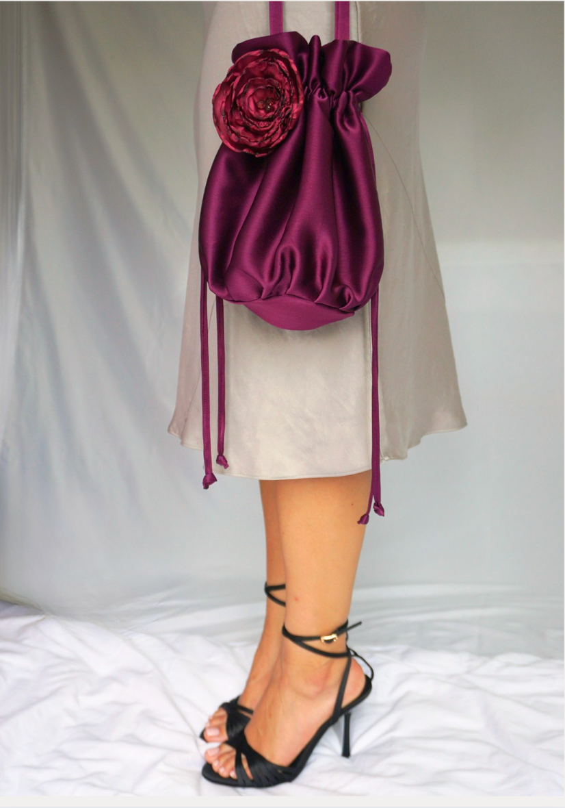 Deep magenta red satin pouch bag with a floral appliqué and drawstring detail, worn with a silver satin dress and black strappy heels.