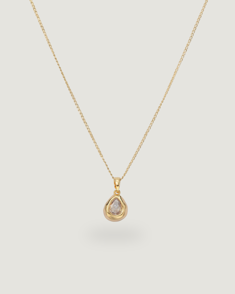 A delicate gold chain necklace with a teardrop-shaped pendant featuring a clear gemstone in a gold bezel setting, displayed against a solid light gray background.