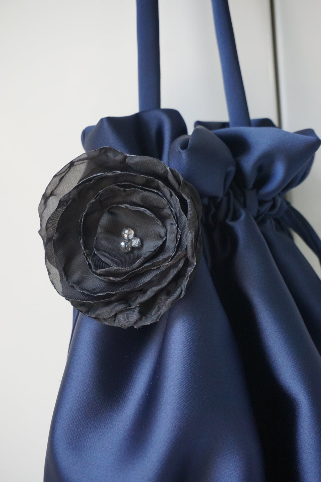 A close-up of a navy blue satin accessory with a large dark gray fabric flower applique featuring a small silver rhinestone center, set against a plain light background.
