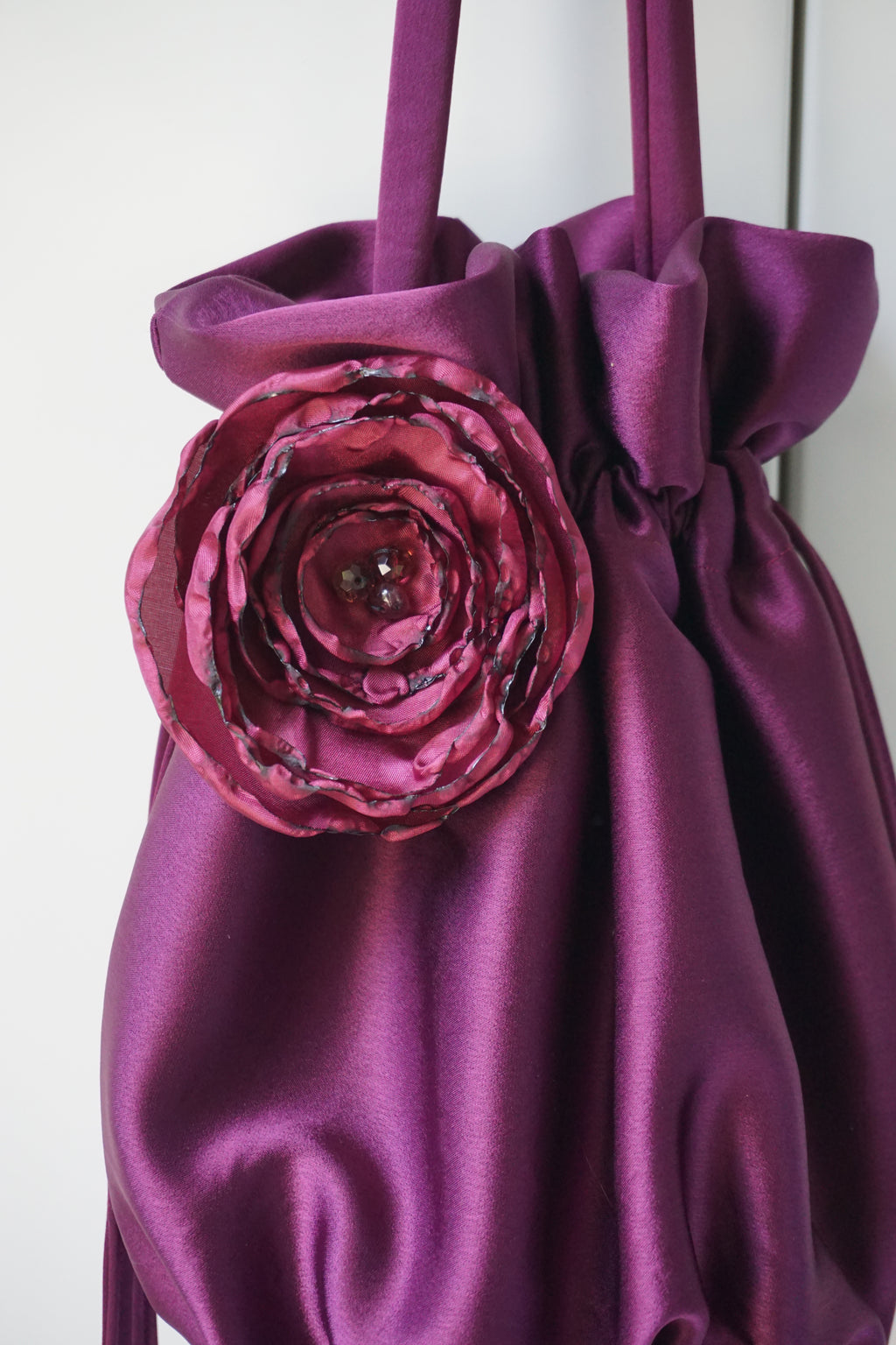 A close-up of a purple satin drawstring bag with a large fabric rose embellishment on the front.