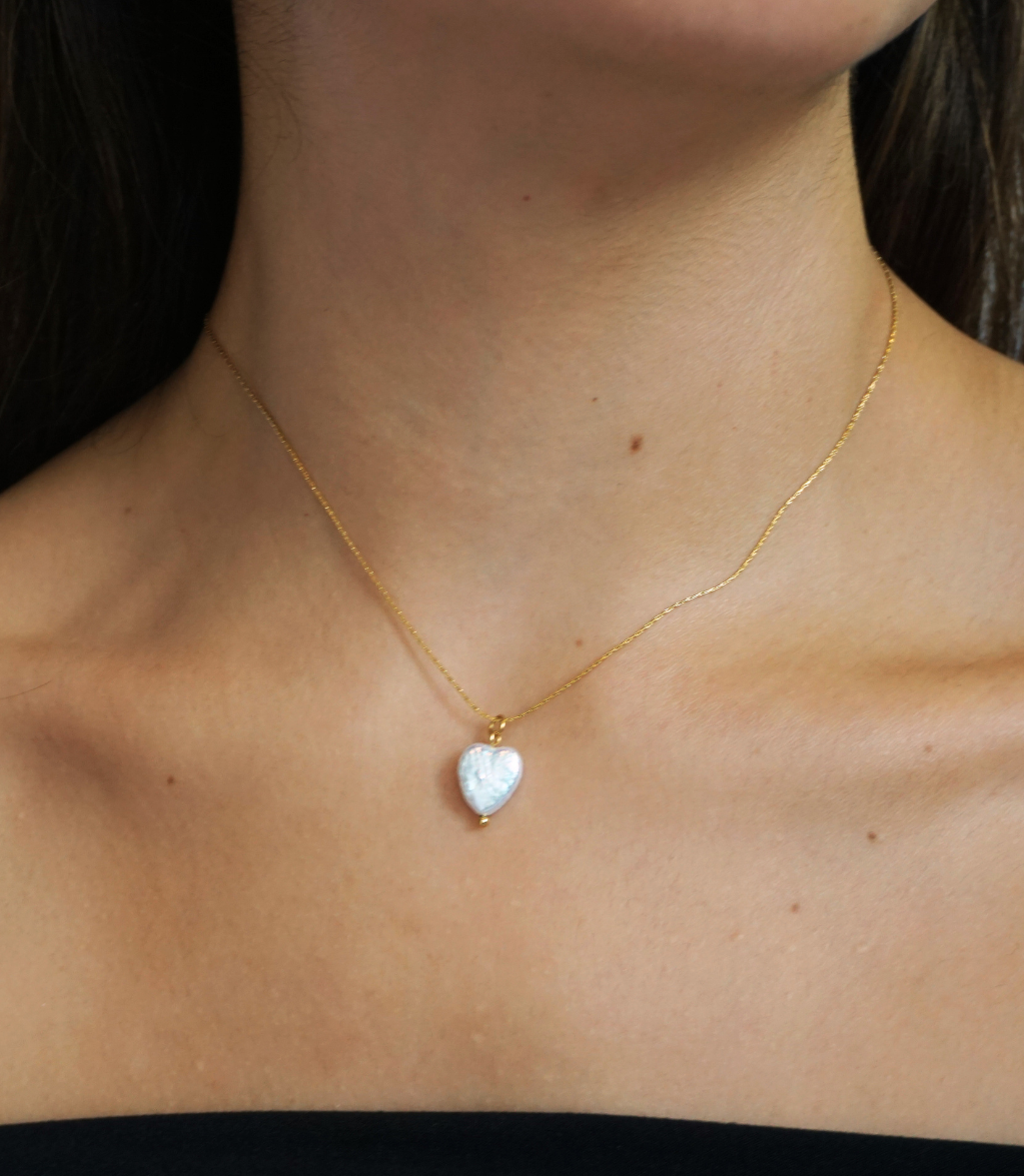 A delicate gold chain necklace with a small, heart-shaped white pearl pendant, shown worn on a model's neck.