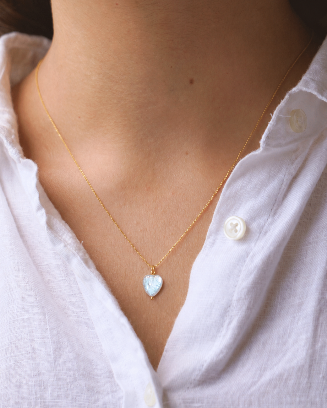A delicate gold chain necklace with a small heart-shaped pendant featuring a light blue iridescent inlay, worn by a model in a white button-down shirt.