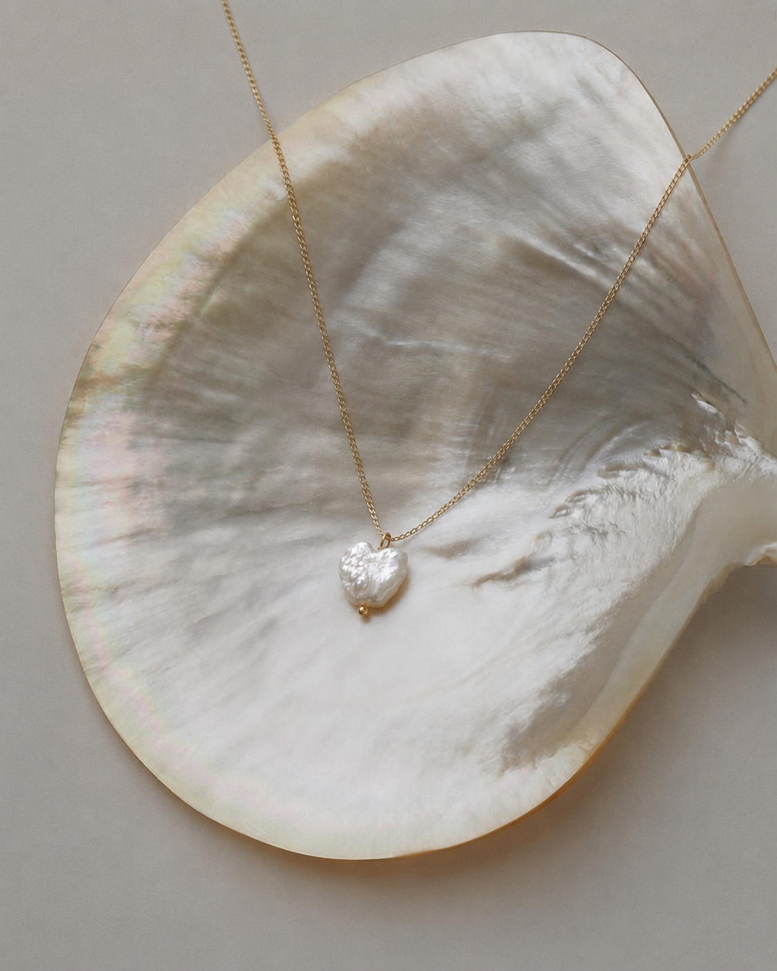 A delicate gold chain necklace with a small, textured white heart-shaped pendant, displayed on a large iridescent mother-of-pearl clam shell against a neutral gray background.