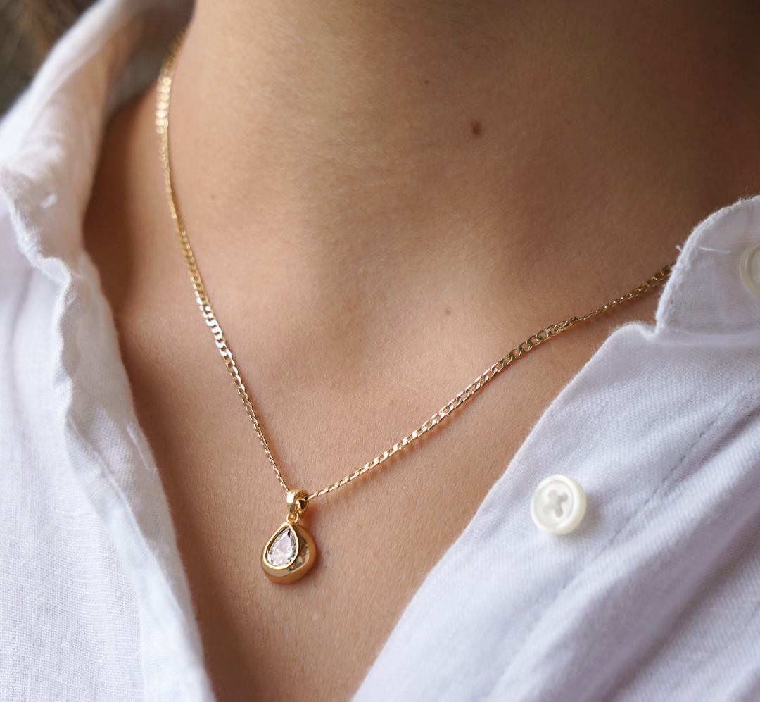 A gold teardrop pendant necklace with a clear stone, worn by a person wearing a white button-down shirt.