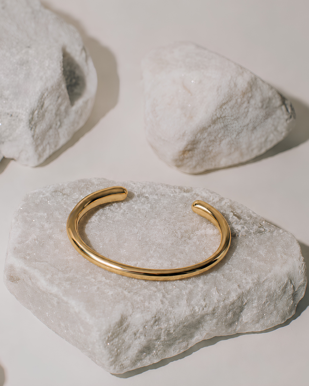 A polished gold open cuff bangle bracelet displayed on a textured white stone, with two more white stones in the background on a light beige surface.