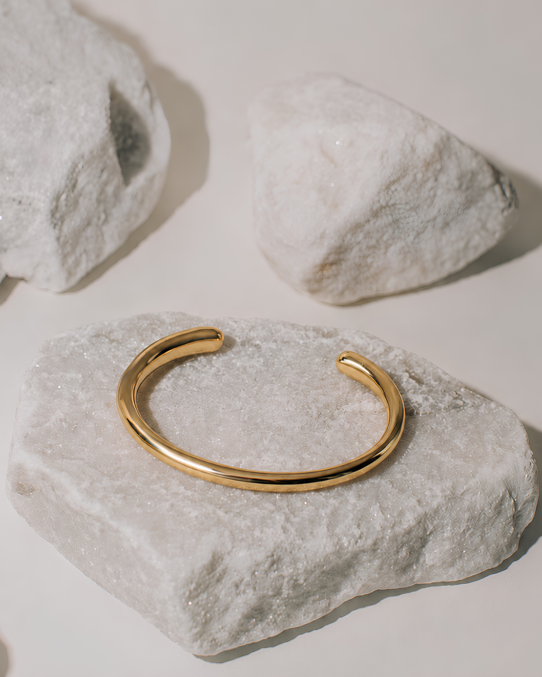 A polished gold open cuff bangle bracelet displayed on a textured white stone, with two more white stones in the background on a light beige surface.