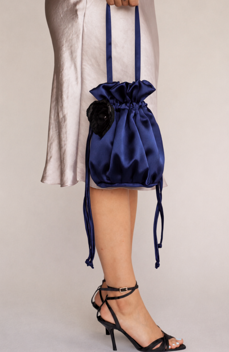 navy blue satin bridal bag with handmade black silk organza flower attached, being held by model wearing a long taupe satin dress wearing black strappy heels on a beige background