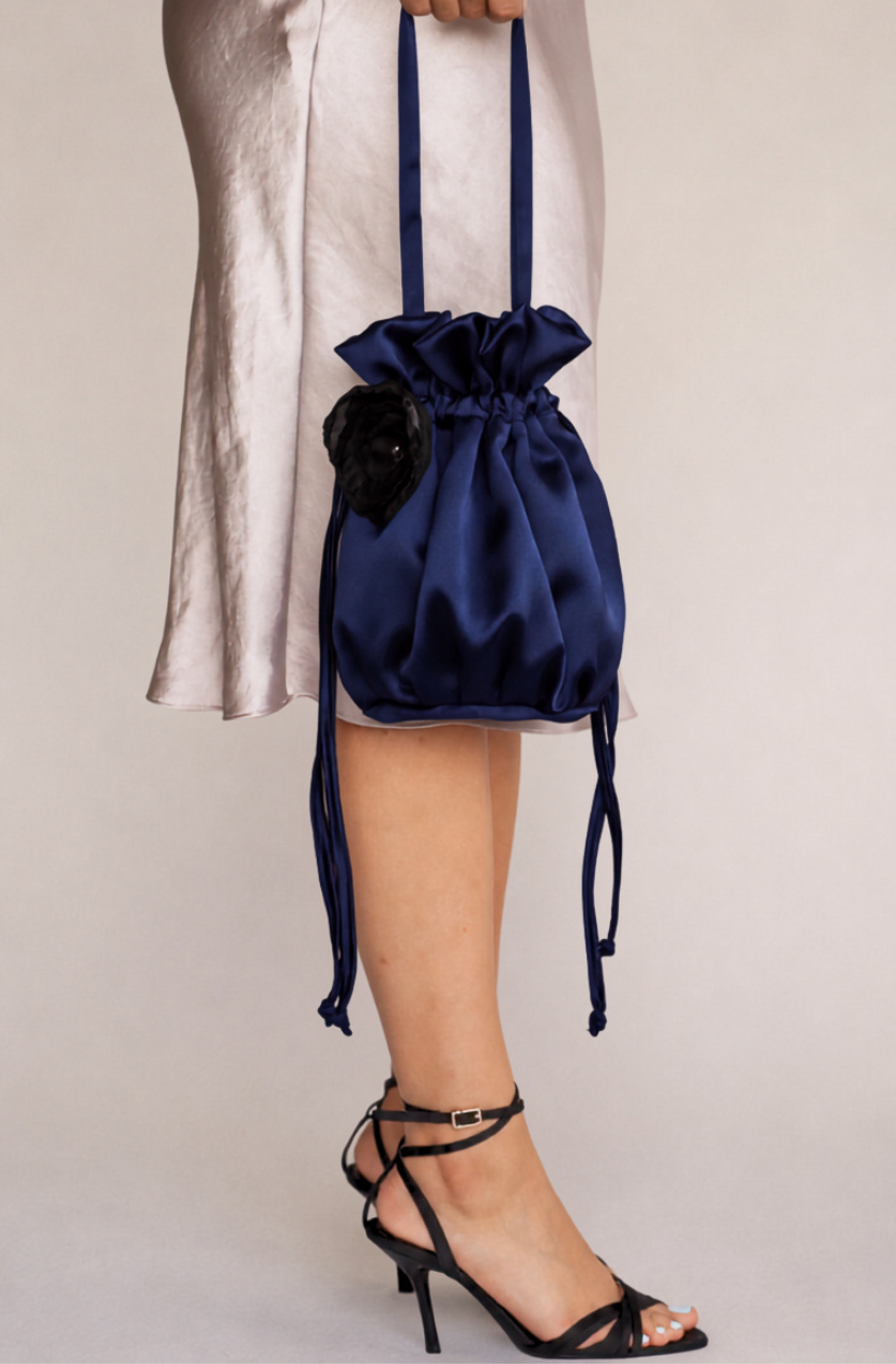 A navy blue satin drawstring bucket bag with a black floral accent, held by a model wearing a light champagne dress and black strappy high-heeled sandals against a neutral beige background.
