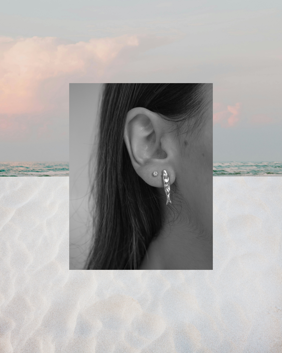 A black and white close-up of a woman's ear wearing a dangling silver earring, centered over a beach scene with pale sand and soft sky.