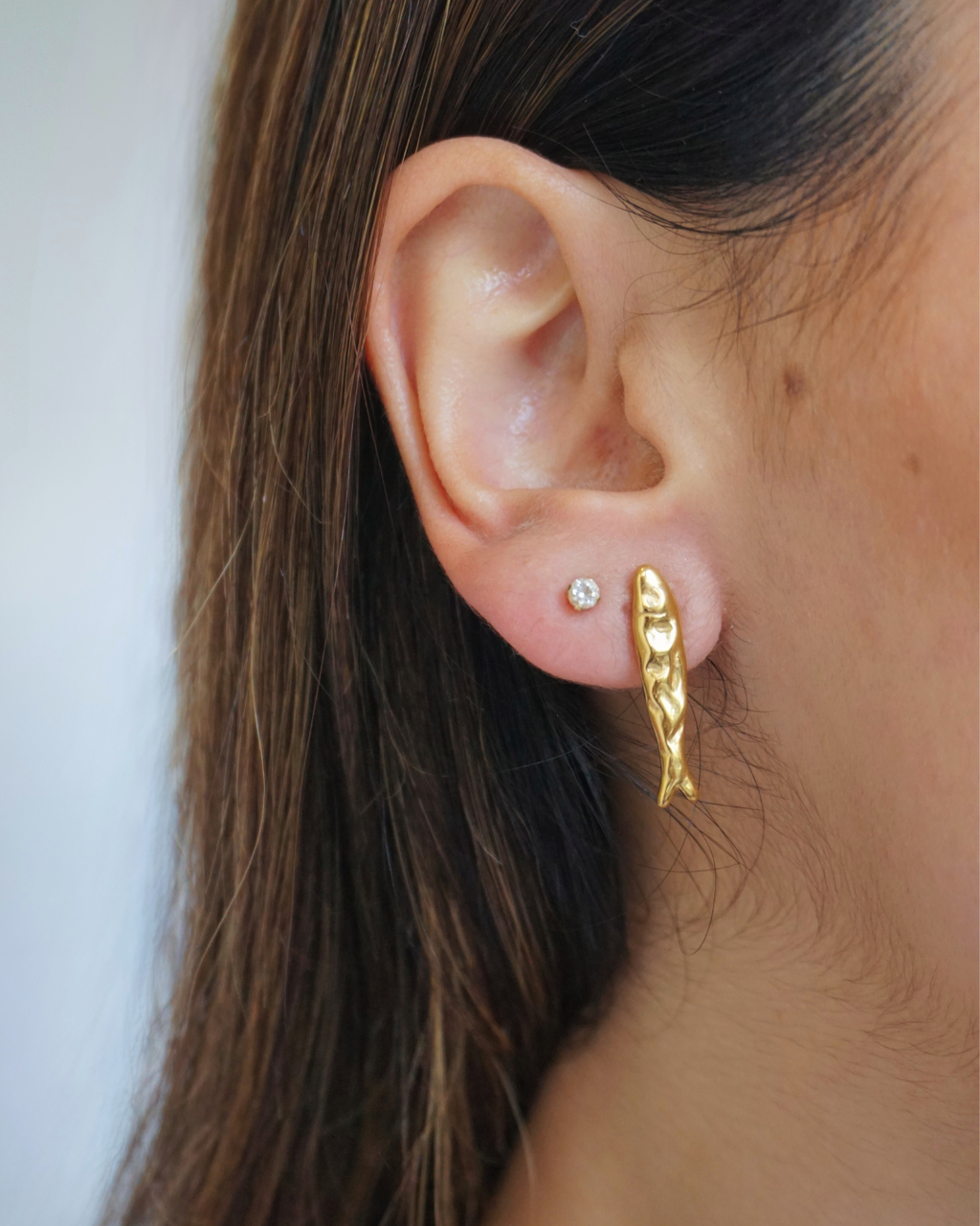 golden fish stud earring on model with brown hair on light beige background