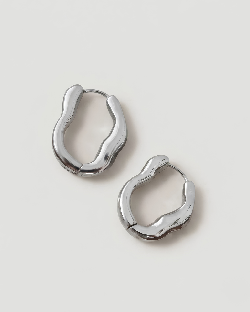 A pair of polished silver huggie hoop earrings with an organic, wavy abstract shape, displayed on a clean light gray background.
