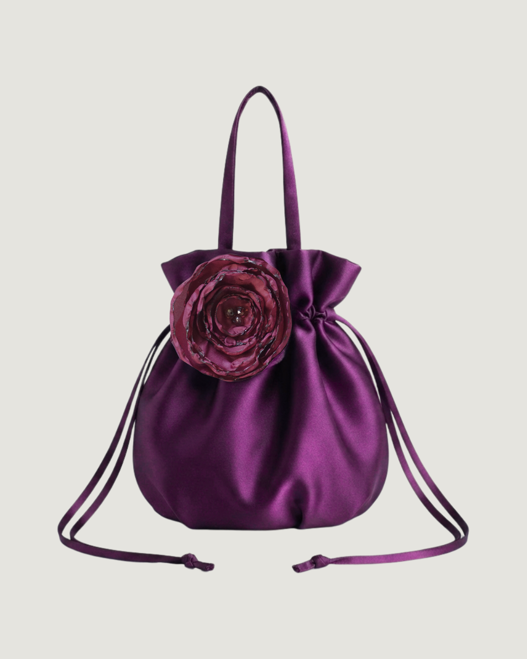A purple satin drawstring bag with a large fabric flower accent on the front, set against a plain light gray background.