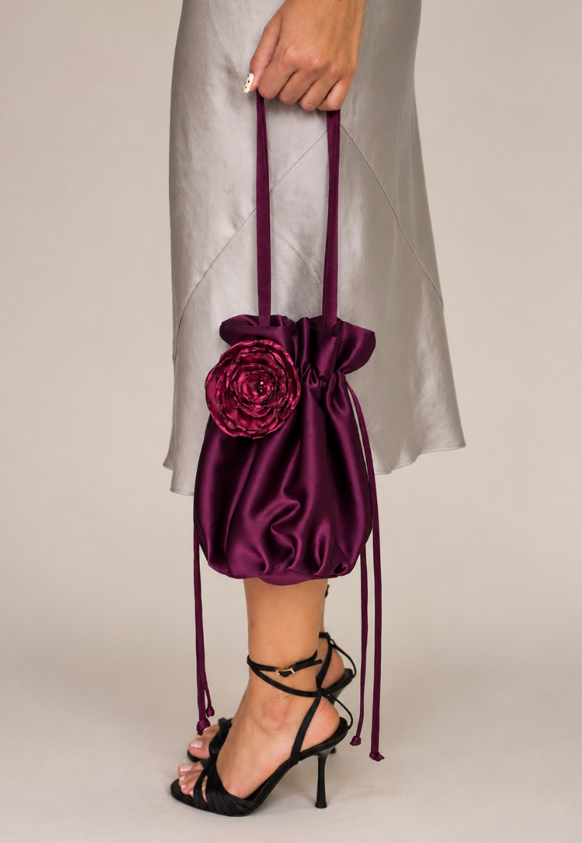 plum satin bridal bag with handmade magenta silk organza flower attached, being held by model wearing a long taupe satin dress wearing black strappy heels on a beige background