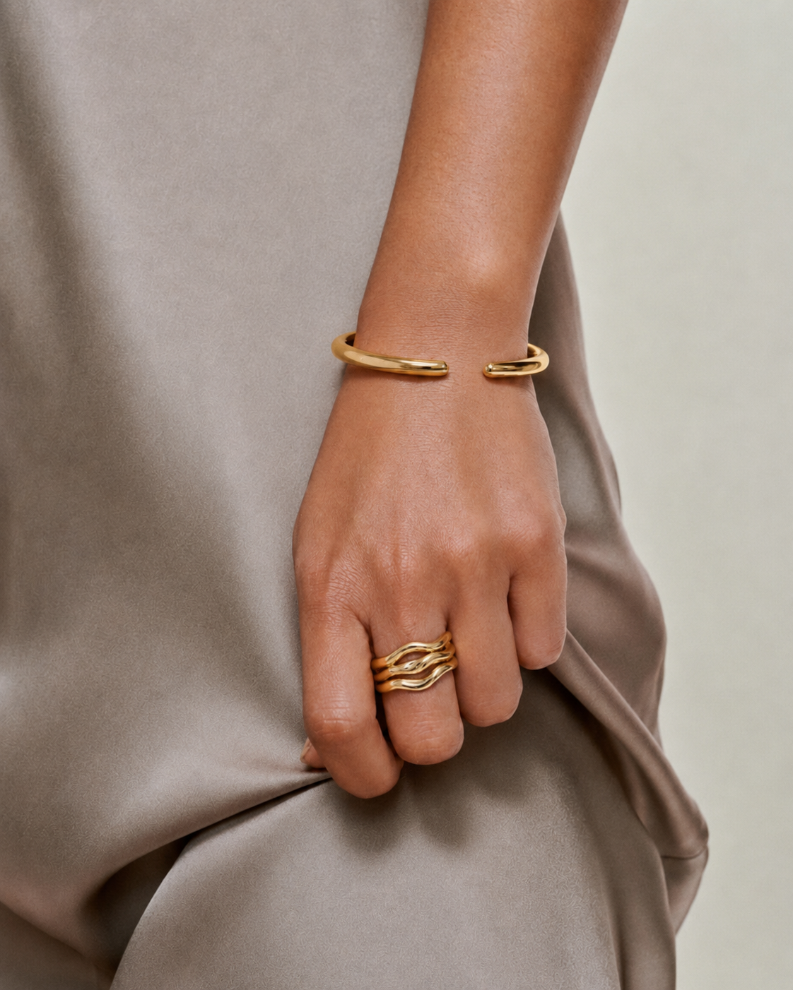 gold cuff bangle and gold ring on models hand, wearing taupe silk wedding guest dress