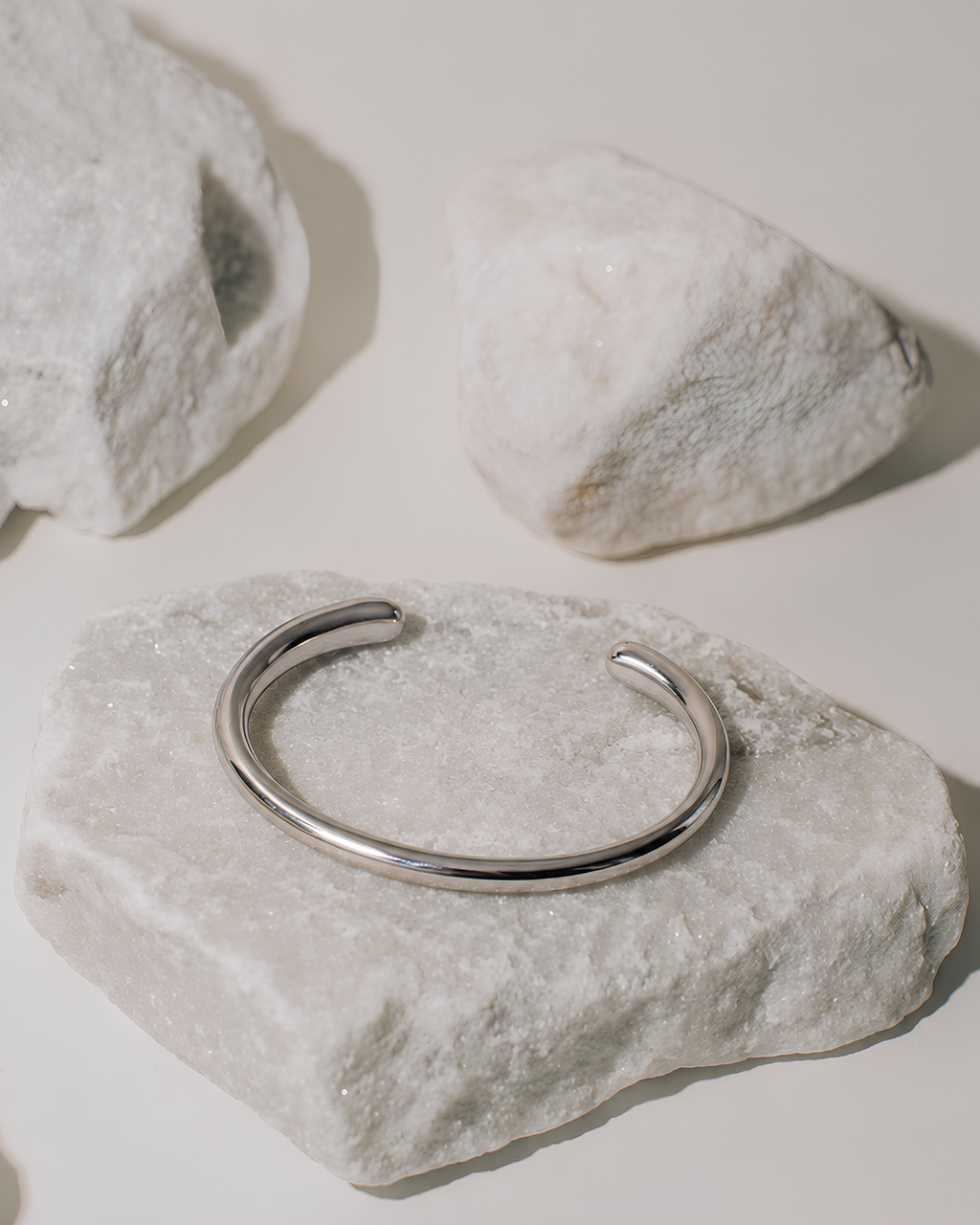 A polished silver open cuff bracelet displayed on a textured white stone, with two additional white stones in the background on a warm off-white surface.