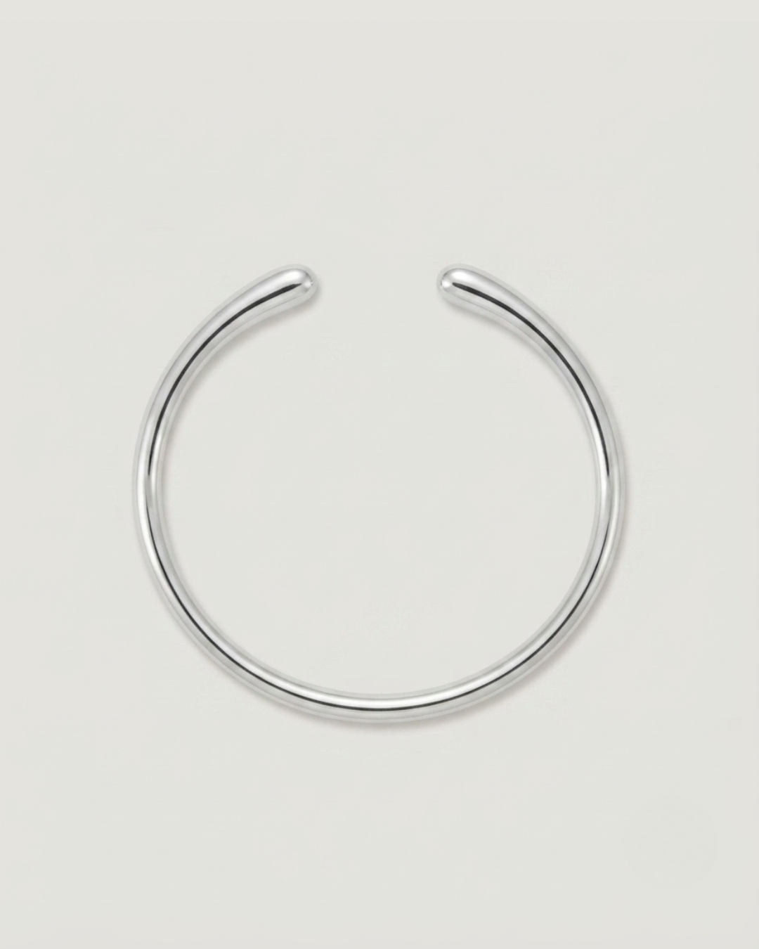 A minimalist silver-toned metal huggie hoop earring with a polished finish, shown on a clean, light gray background.