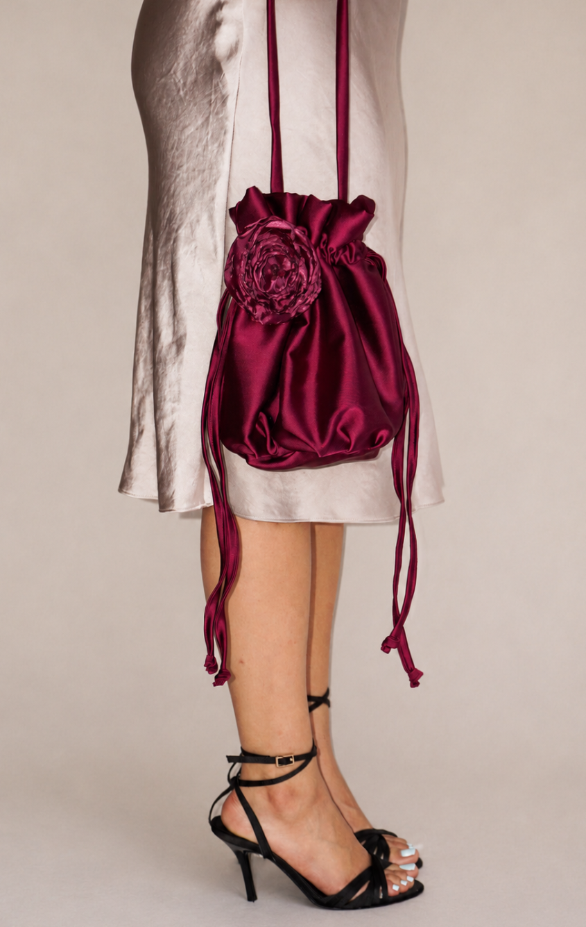 A burgundy satin drawstring crossbody bag with a fabric flower accent, shown worn by a model in a cream satin dress and black strappy heels against a beige studio background.
